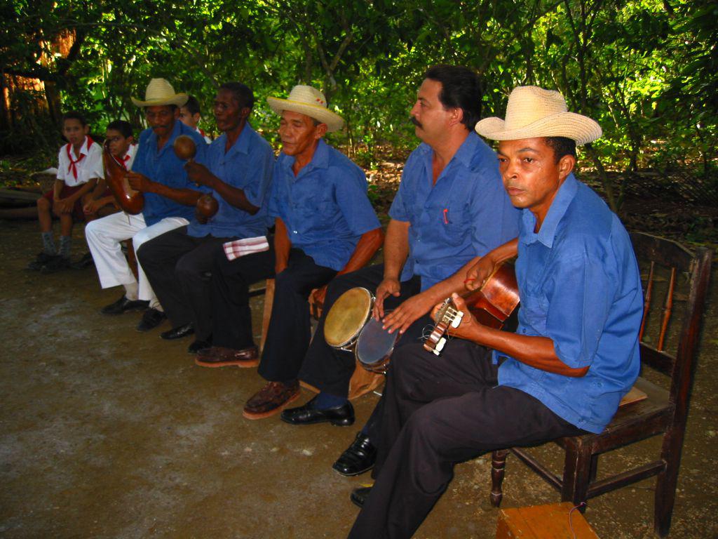 Día. Exterior. Primer plano de grupo musical sentado, tocando. Niños al fondo. Vegetación detrás.