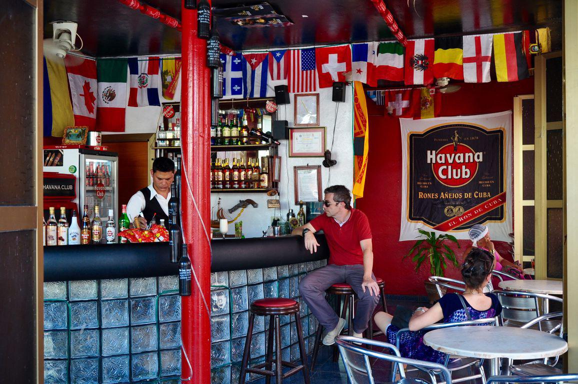 Día. Exterior. Vista de la barra del bar Havana Club con cantinero y hombre sentado a la barra. Mujer sentada de lado